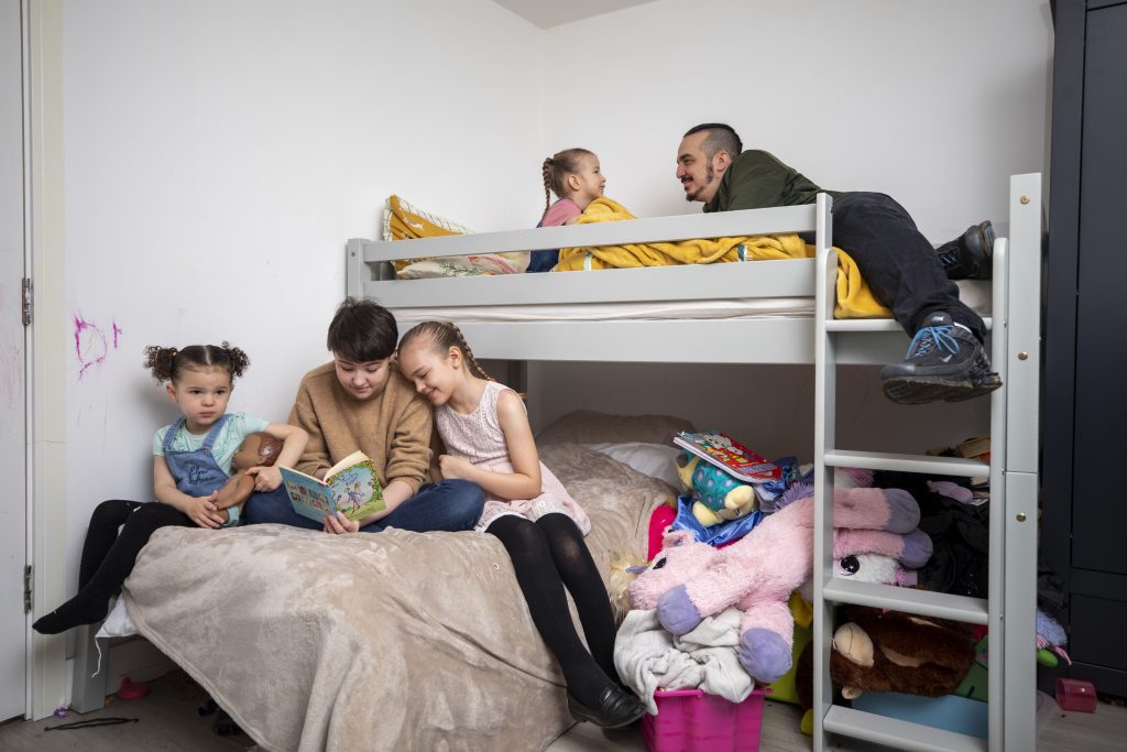 Damien reading to one of his daughters while the others also read on the 22nd of January 2022 in the Medway region of Kent. The family are currently living in temporary accommodation and have been receiving support from HomeStart Medway. (photo by Andy Aitchison / HomeStart UK)